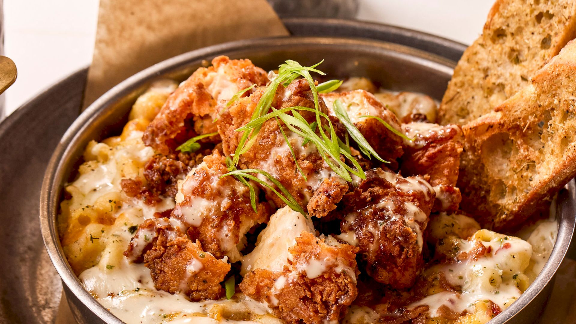 A bowl of creamy mac and cheese topped with crispy chicken and garnished with herbs, served with bread slices; popcorn in the background.