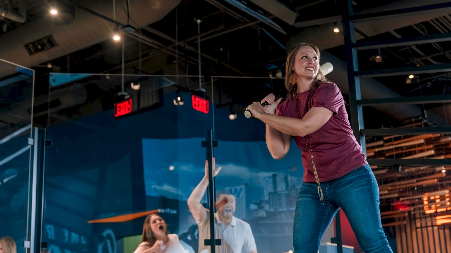 A person is playing an interactive baseball simulation in a fun, indoor sports-themed venue with a small group of onlookers watching.