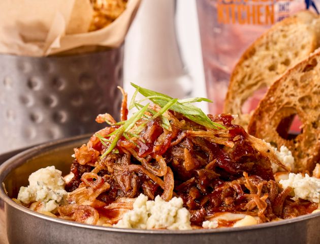 The image shows a dish with pulled pork, crispy onions, blue cheese, and bread, with peanuts and a drink in the background.