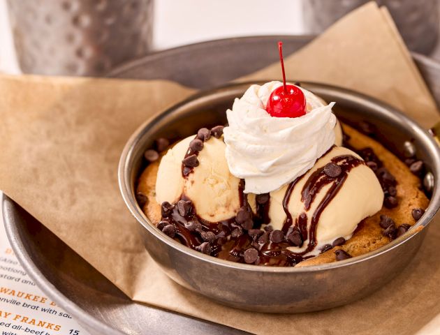 A skillet dessert with ice cream, whipped cream, chocolate sauce, and a cherry on top sitting on parchment paper.