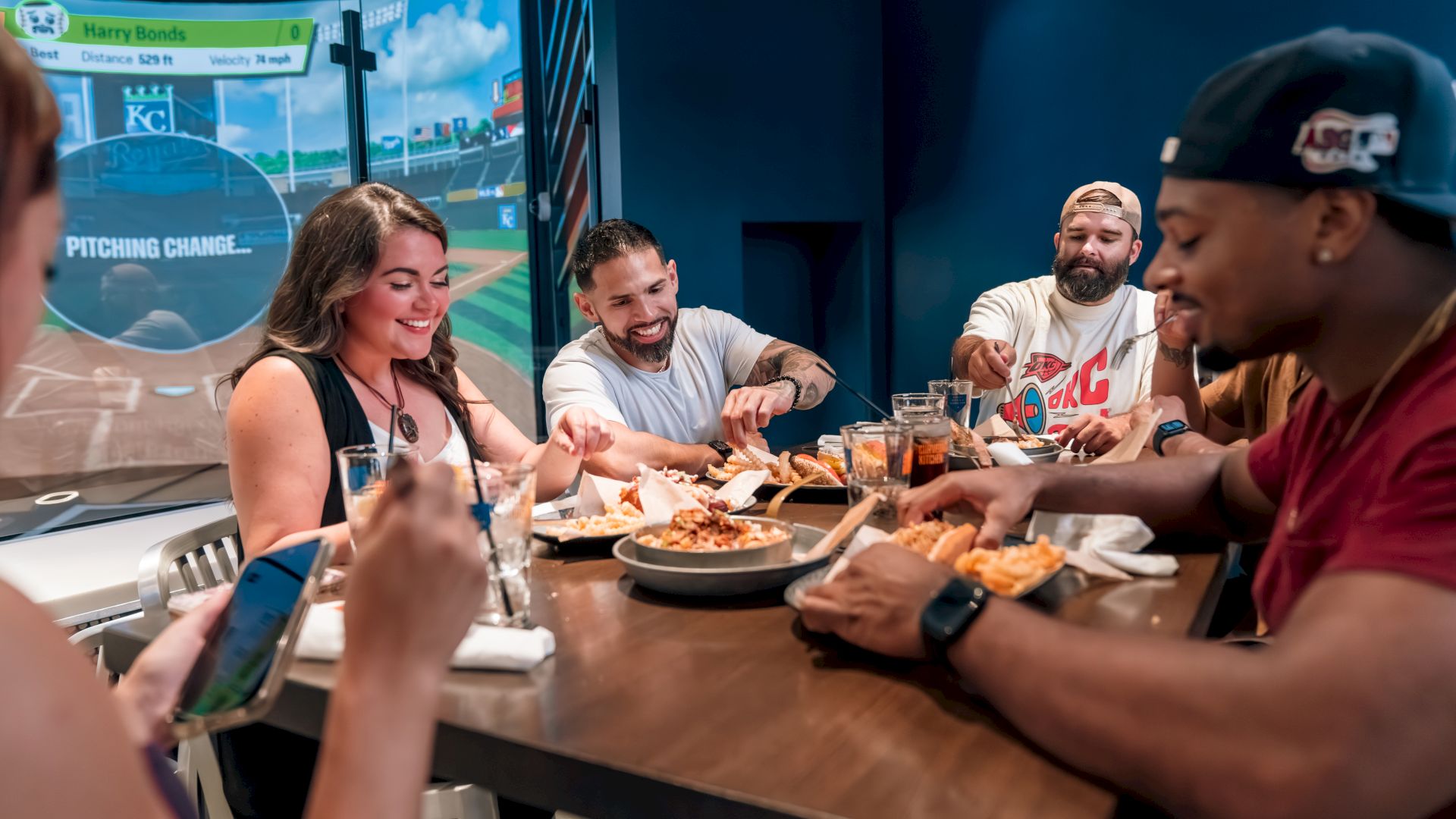 A group of people is sitting around a table, enjoying food and drinks in a lively setting with a large screen showing a baseball field.