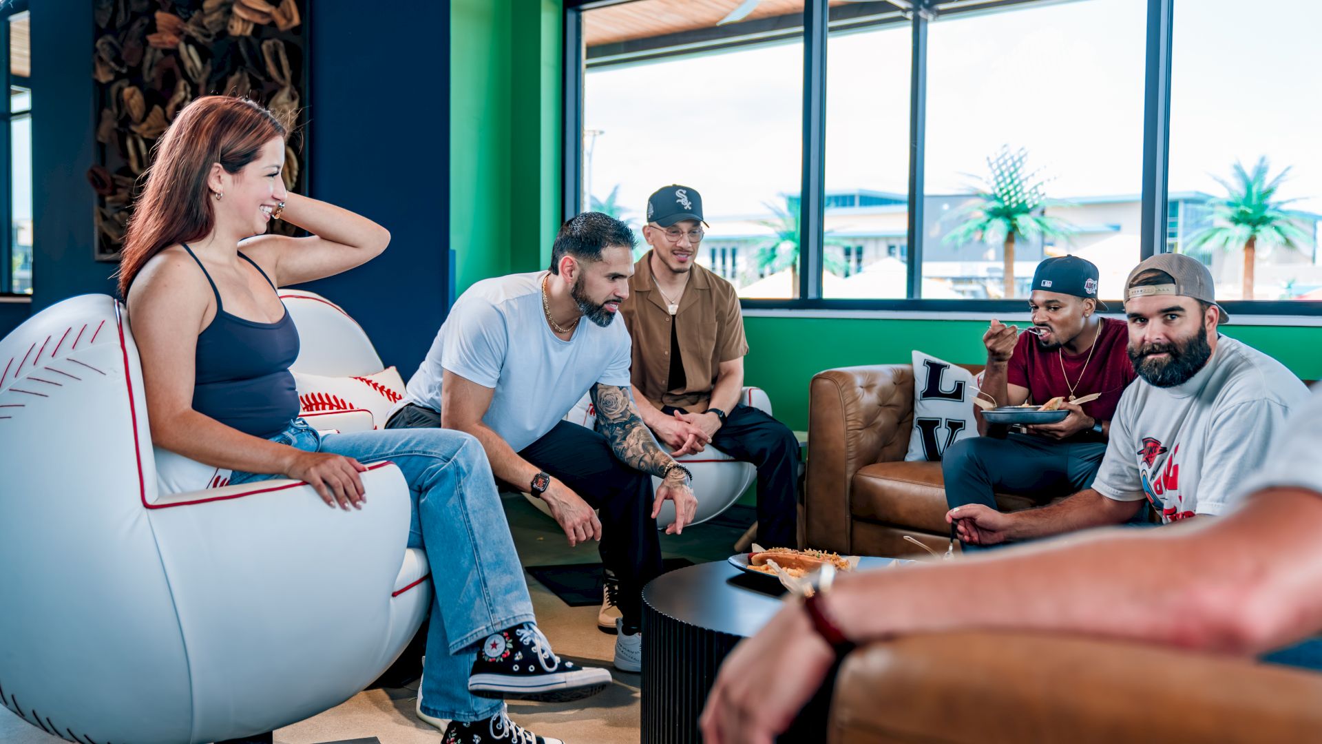 A group of people are sitting and talking in a casual indoor setting with colorful decor and large windows.