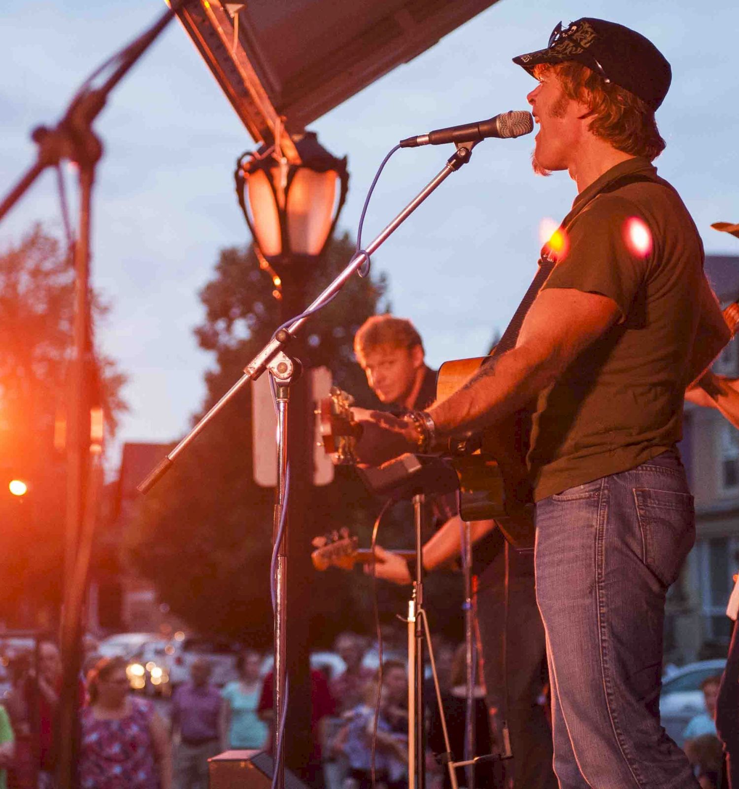 A live band playing on stage outdoors at sunset with guitars, a vocalist at a mic, and a small crowd watching. Top it at 140 characters, ending with a period.