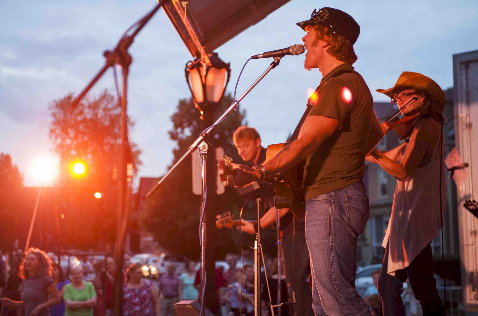 A band is performing on an outdoor stage at dusk with a crowd watching, illuminated by stage lights.