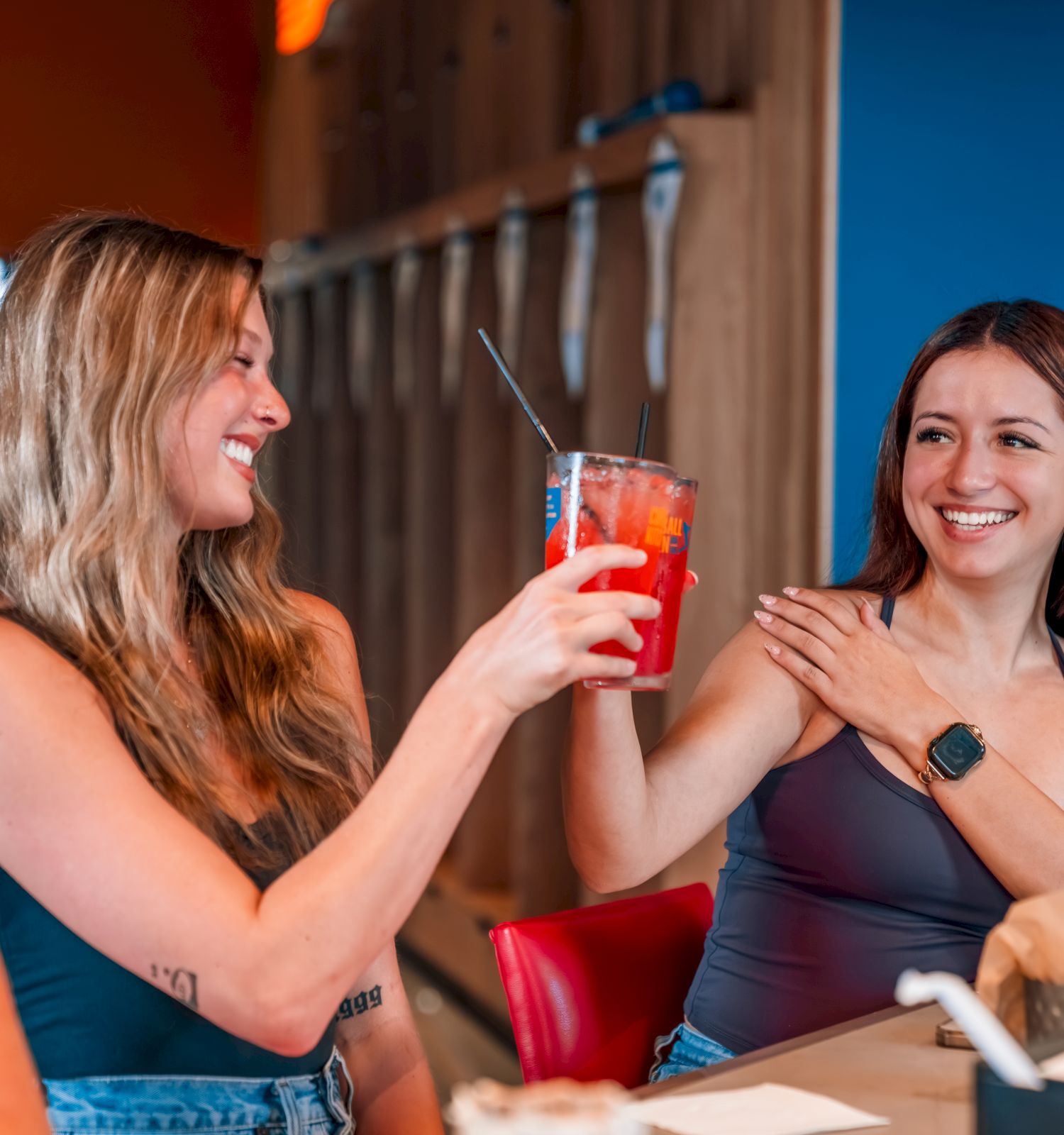 Two friends laugh and clink bright red drinks at a caf&eacute; table, sharing a fun moment together.