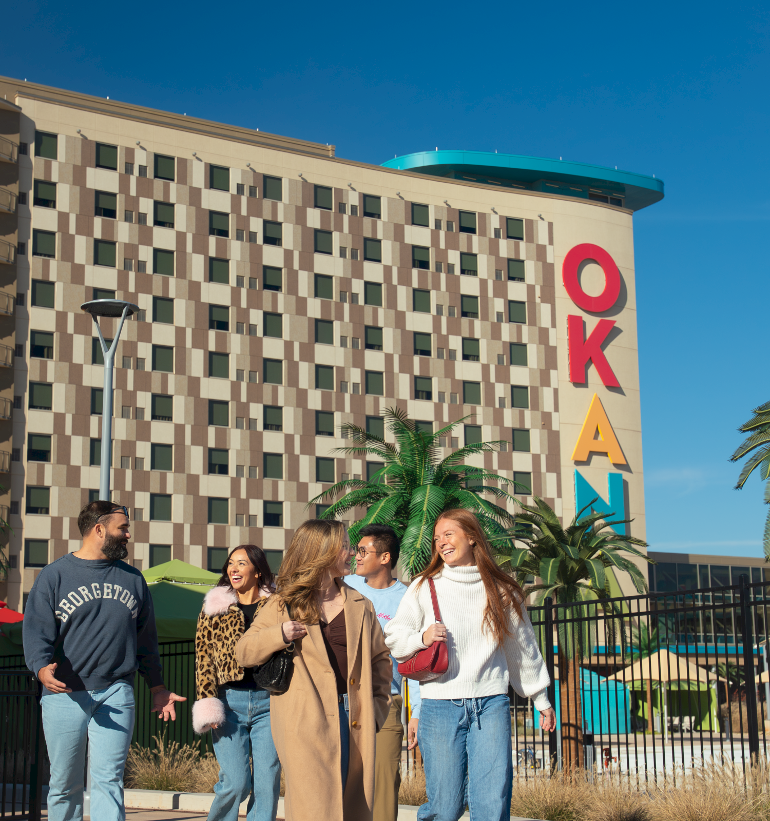 A group of students walk outside a campus building with colorful &ldquo;OKA&rdquo; letters under a clear blue sky.