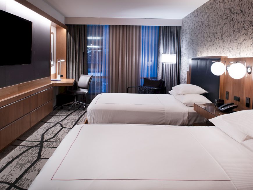 A modern hotel room with two beds, a flat-screen TV, a desk with a chair, and stylish lighting and decor, creating a cozy atmosphere.