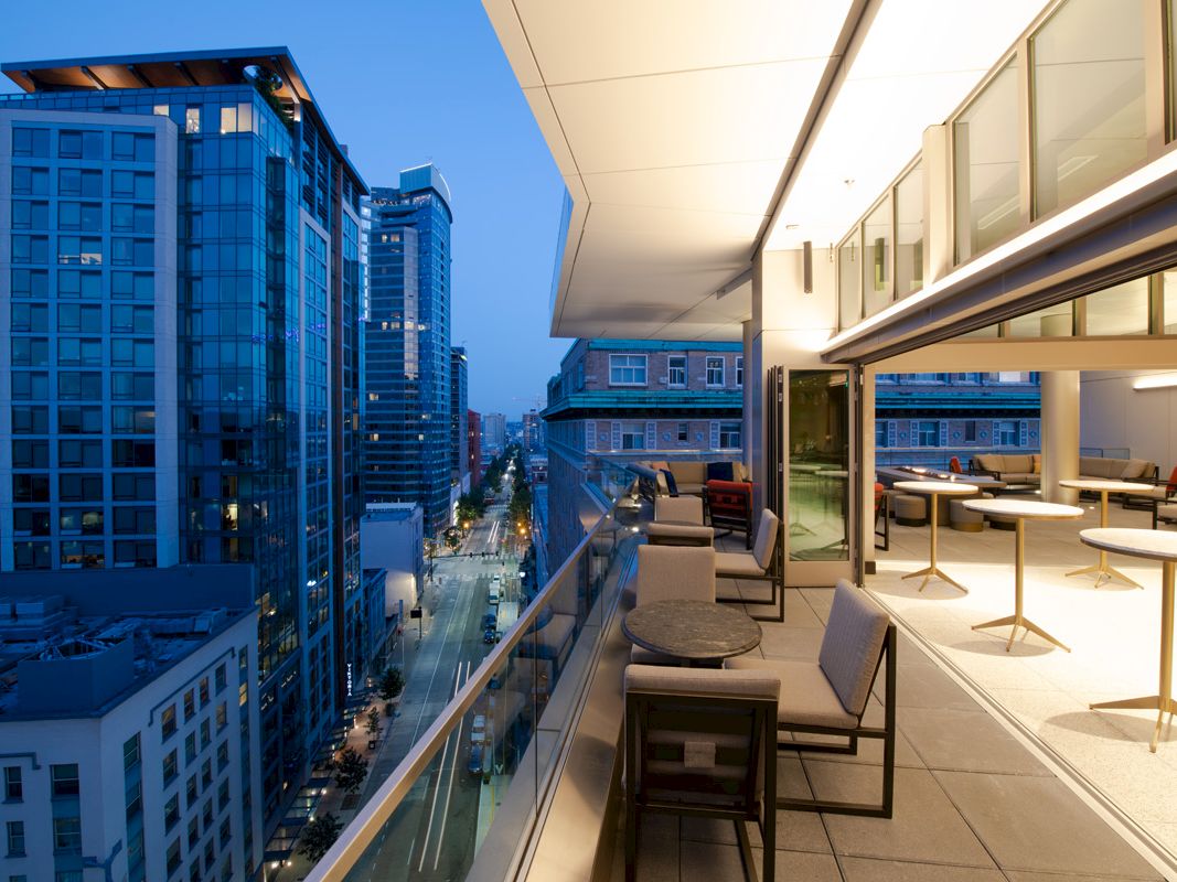 A modern urban balcony with seating overlooks city buildings and streets at dusk, with a sleek interior visible through open doors.