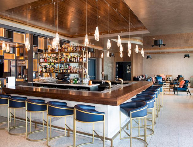 The image shows a modern bar with a U-shaped wooden counter, blue barstools, hanging lights, and a well-stocked shelf of bottles in the background.