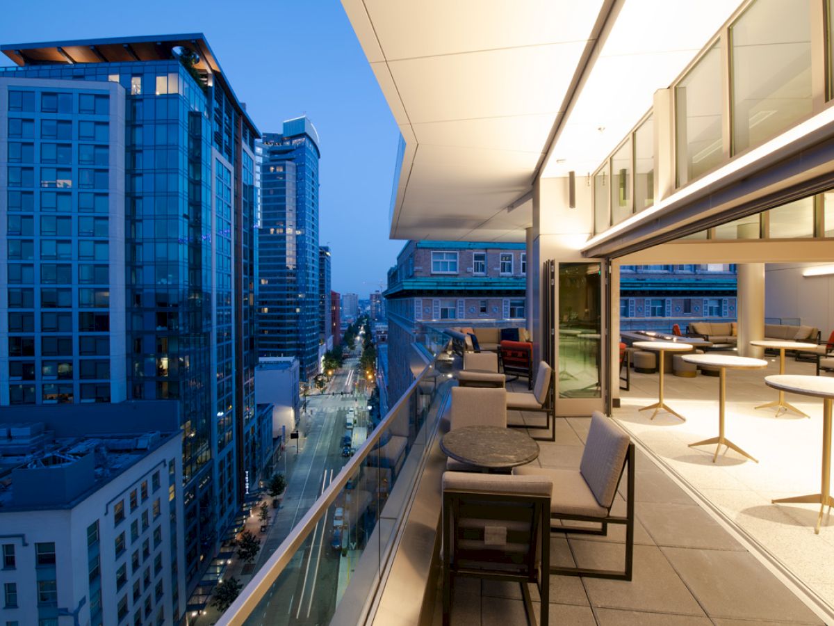 A modern cityscape view from a high balcony with outdoor seating and tables, overlooking illuminated skyscrapers at dusk.