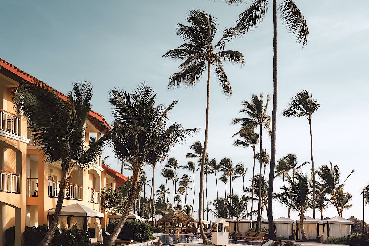 A luxurious poolside scene with lounge chairs, palm trees, and a building, set against a clear blue sky.
