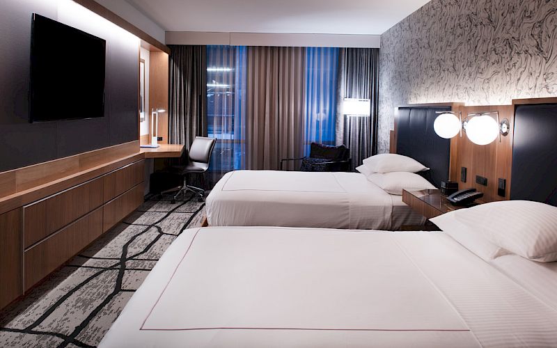 The image shows a modern hotel room with two beds, a TV, desk, and stylish decor.